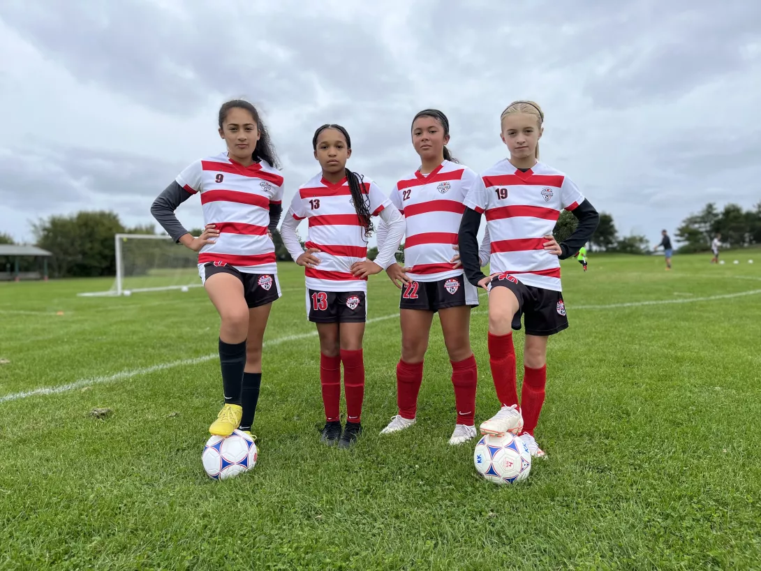 Four girls standing together on the field posing for a picture with soccer balls